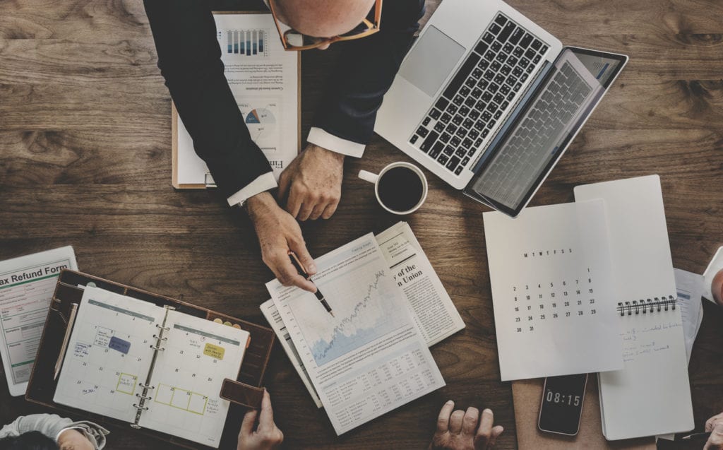Overhead view of a business meeting with hands analyzing financial reports, charts, and a laptop, emphasizing investment strategies in the medical device sector.